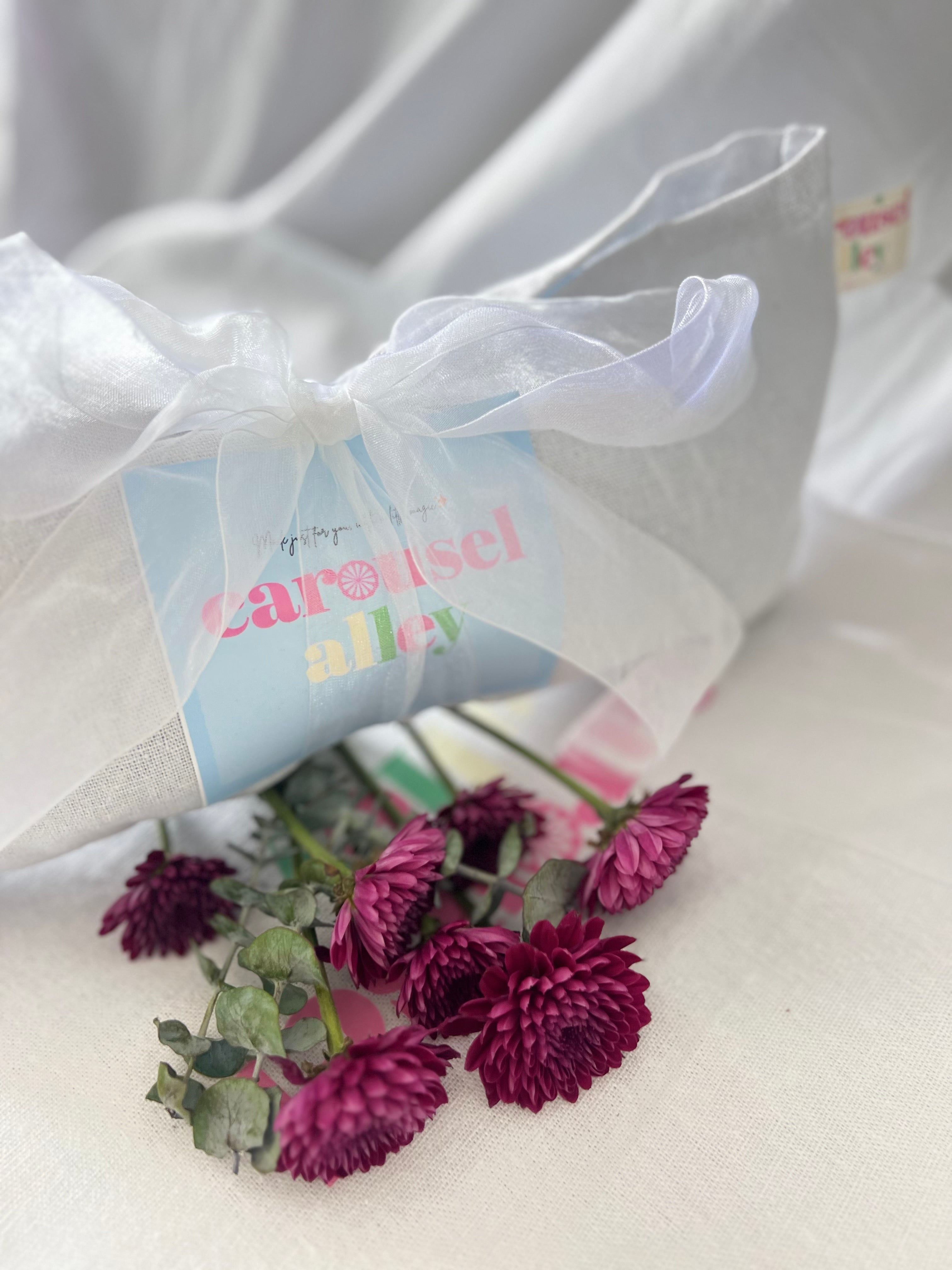 Bouquet of purple flowers with a 'Carousel Alley' gift bag on a white background. Linen keepsake bag 